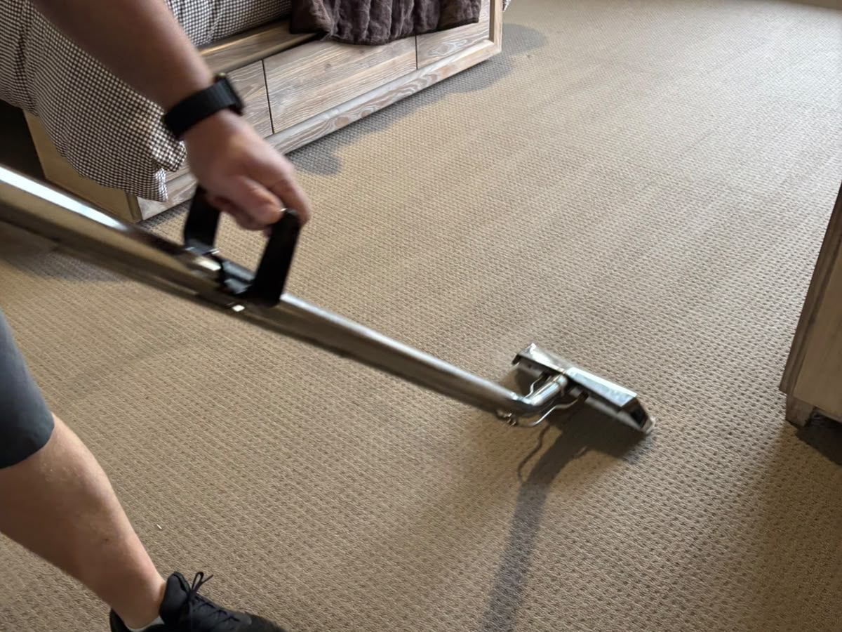 Operator guiding a stainless steel carpet cleaning wand across pale carpet beside a bed