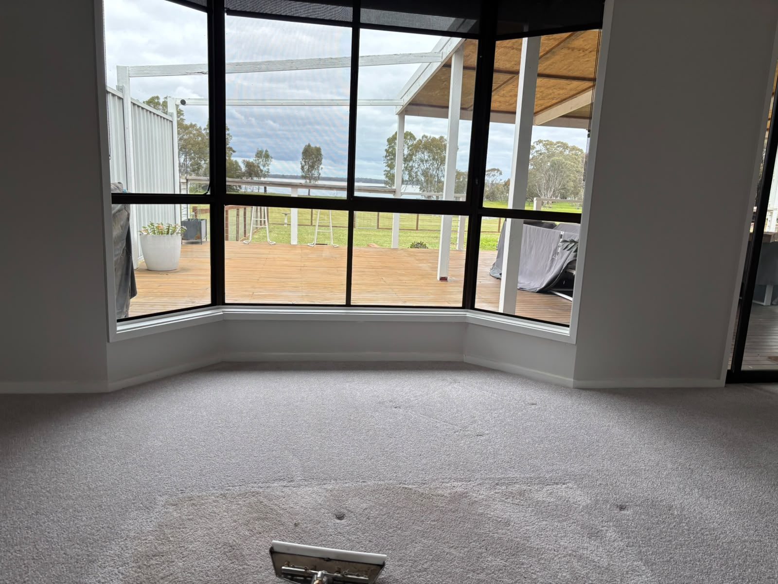 Grey carpet in a lounge room with a bay window looking out to a timber deck, with a cleaning wand head at the edge of the frame